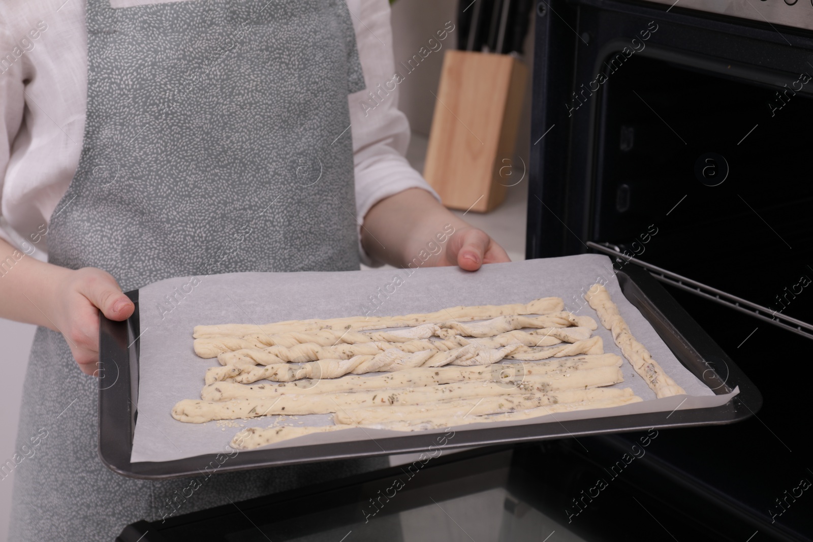 Photo of Woman holding baking sheet with homemade breadsticks near oven in kitchen, closeup. Cooking traditional grissini