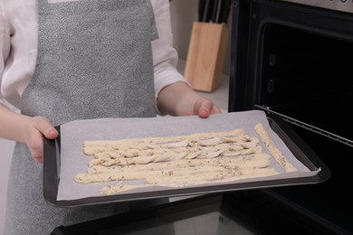 Photo of Woman holding baking sheet with homemade breadsticks near oven in kitchen, closeup. Cooking traditional grissini
