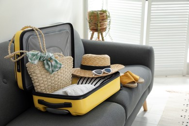 Photo of Open suitcase full of clothes, shoes and summer accessories on sofa in room