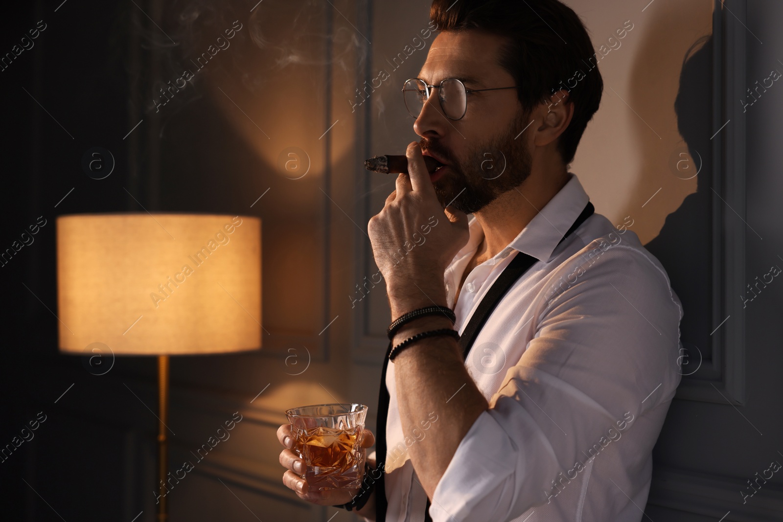 Photo of Handsome man with glass of whiskey smoking cigar at home. Space for text