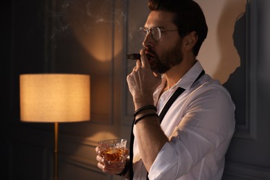 Photo of Handsome man with glass of whiskey smoking cigar at home. Space for text