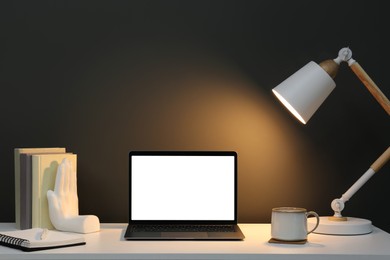 Photo of Stylish workplace with laptop, lamp, cup and decor on white table near grey wall