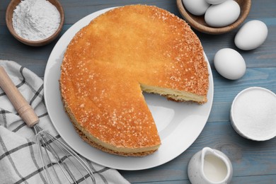 Tasty sponge cake, whisk and ingredients on light blue wooden table, flat lay