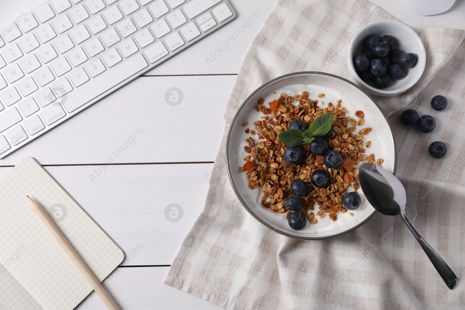 Photo of Delicious granola with blueberries in bowl, stationery and computer keyboard on white wooden table, flat lay