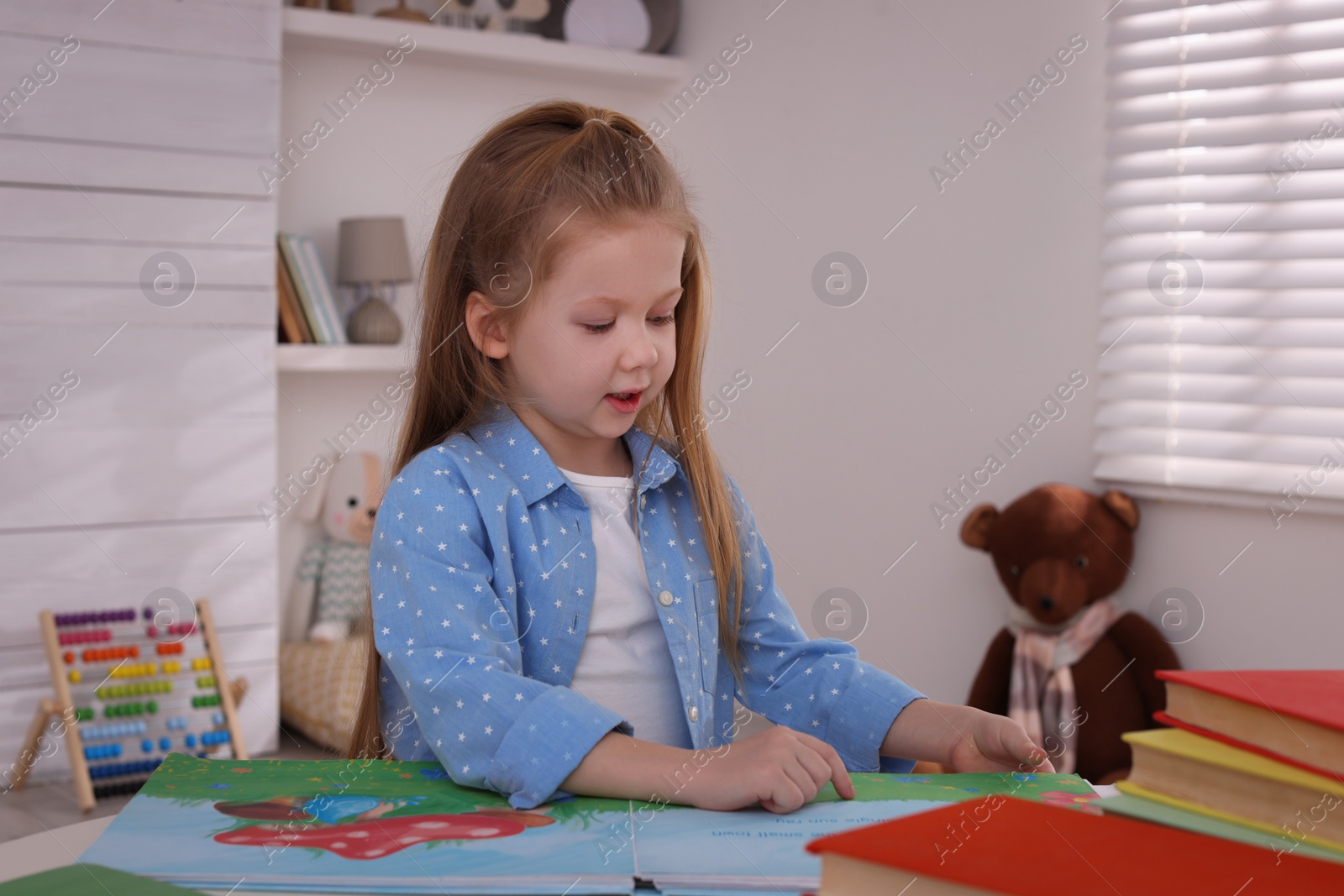 Photo of Cute little girl reading book at table in room