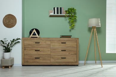 Photo of New wooden chest of drawers, plants and lamp near green wall in stylish room