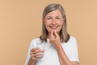 Senior woman with glass of water taking pill on beige background