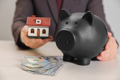 Man holding house model, piggy bank and dollar banknotes at wooden table, closeup. Saving money concept