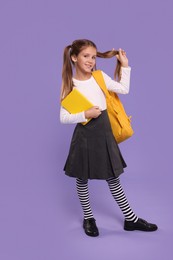 Smiling schoolgirl with backpack and book on violet background