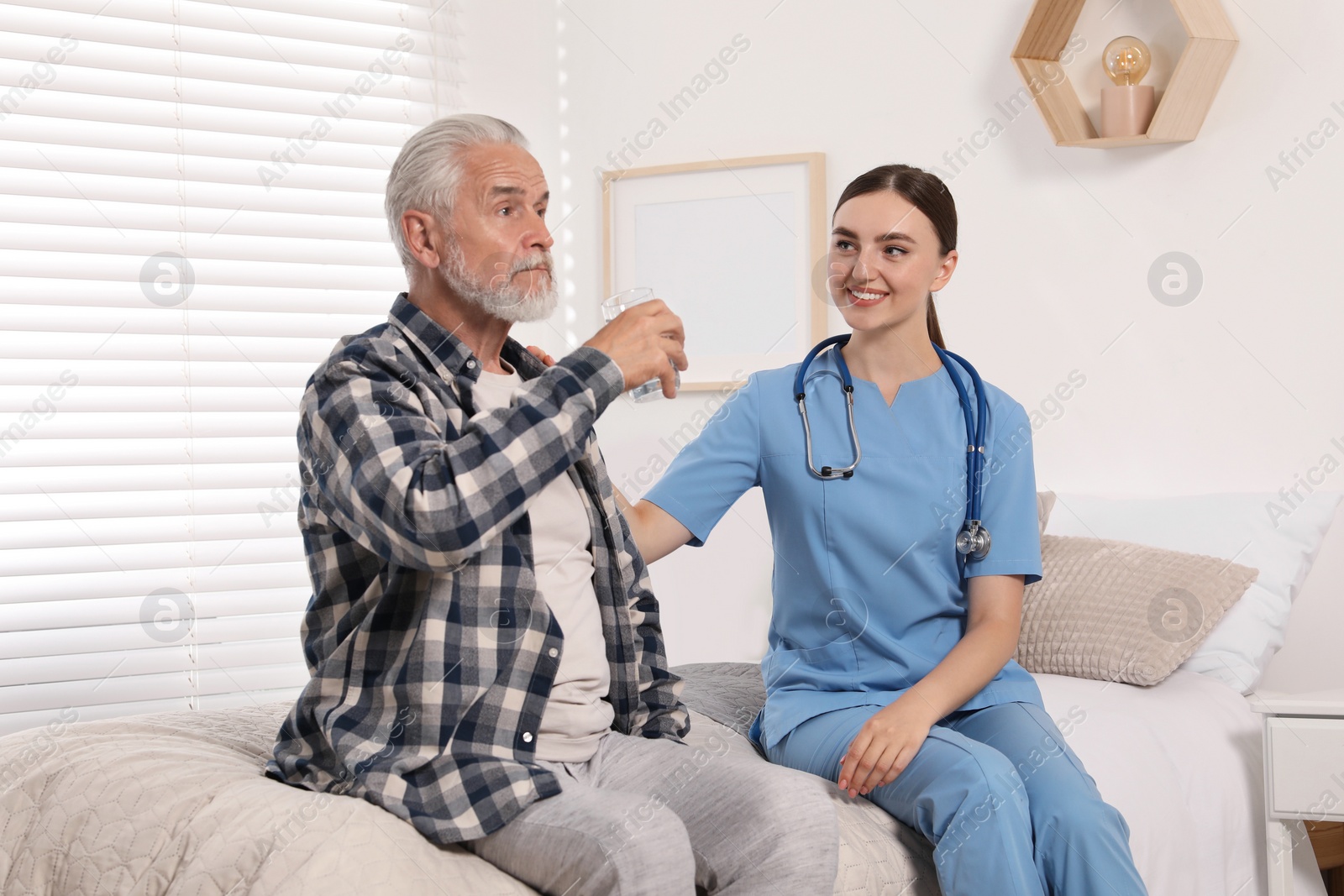 Photo of Senior man with glass of water and young healthcare worker sitting on bed indoors