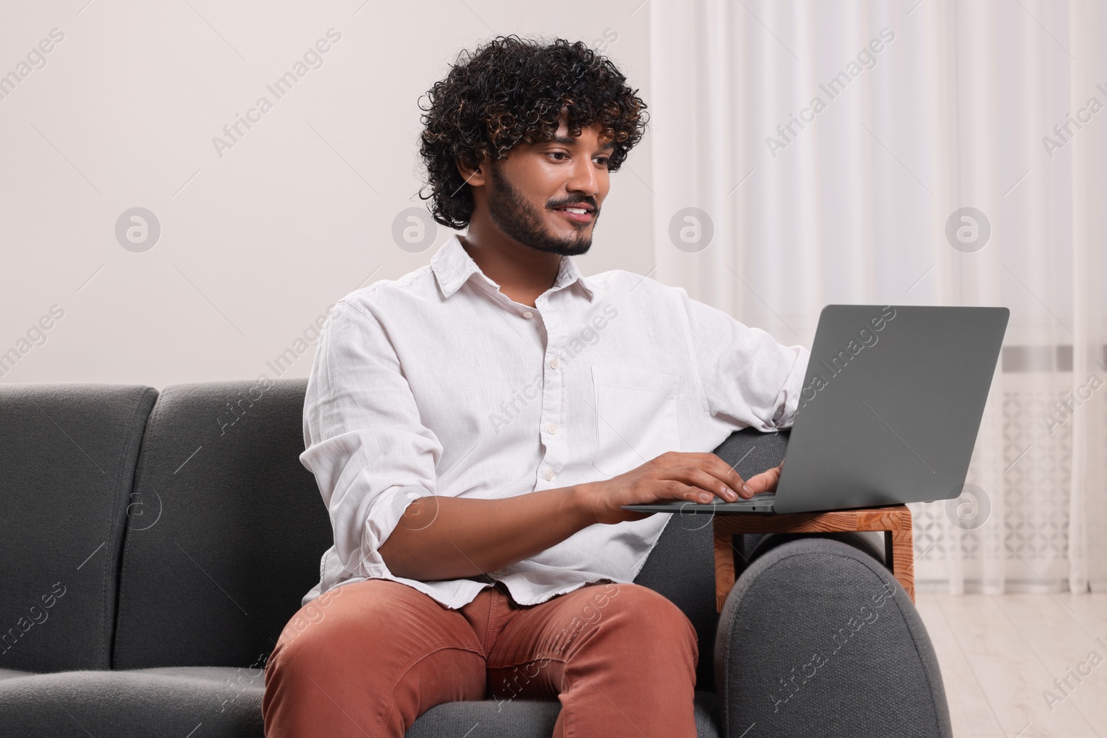 Photo of Happy man using laptop on sofa with wooden armrest table at home