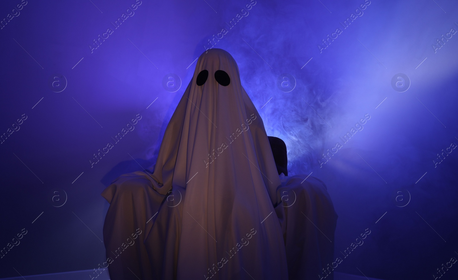 Photo of Creepy ghost. Woman covered with sheet sitting in armchair in blue light