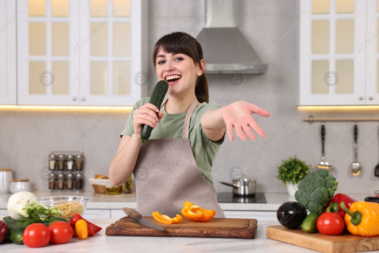 Photo of Happy young housewife with fresh cucumber having fun while cooking at white marble table in kitchen