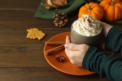 Photo of Woman holding cup of tasty pumpkin spice latte with whipped cream at wooden table, closeup. Space for text
