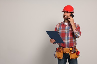 Professional builder in hard hat with clipboard and tool belt talking on phone against light background, space for text