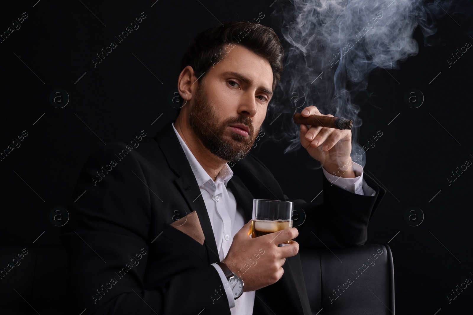 Photo of Handsome man in elegant suit with glass of whiskey smoking cigar on black background