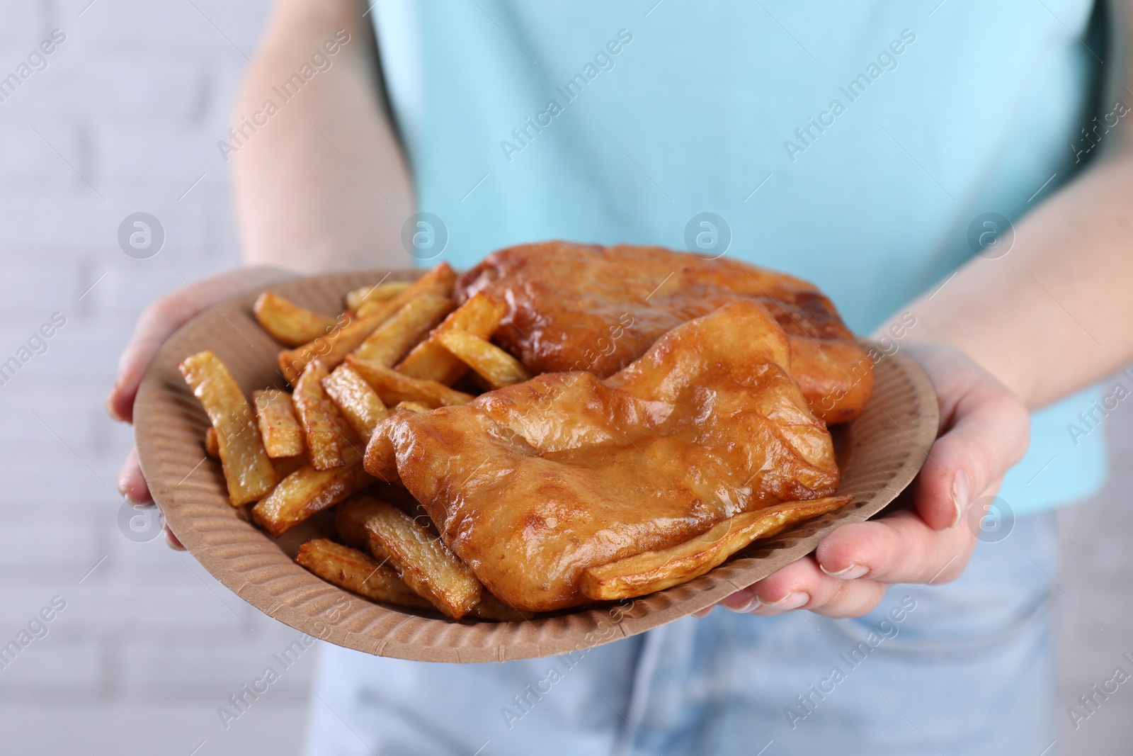 Photo of Woman holding fish and chips in paper plate on near white brick wall, closeup