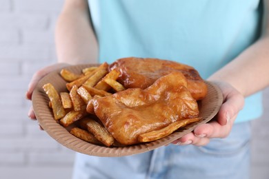 Woman holding fish and chips in paper plate on near white brick wall, closeup