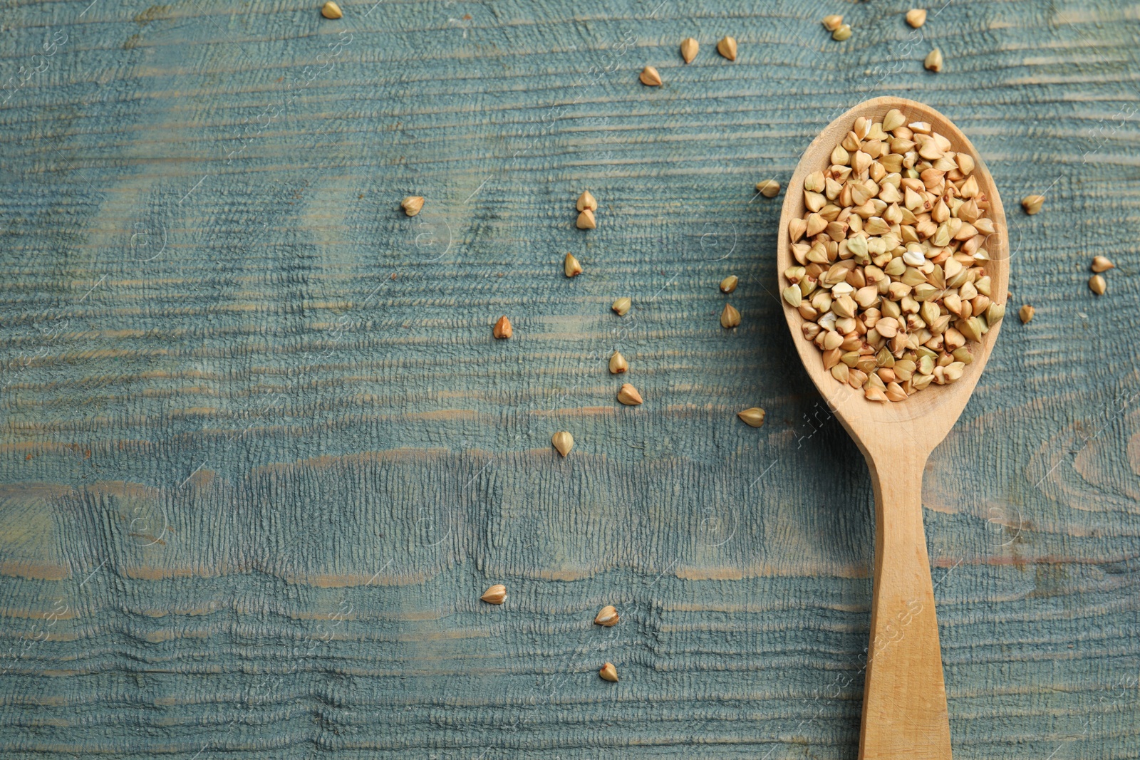 Photo of Uncooked green buckwheat grains in spoon on light blue wooden table, top view. Space for text