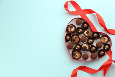 Different delicious chocolate candies in heart shaped box and ribbon on light blue background, top view. Space for text