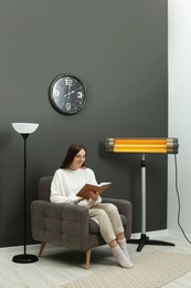 Photo of Young woman reading book on armchair near electric infrared heater at home