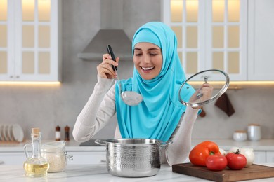 Photo of Muslim woman making delicious soup with vegetables at white table in kitchen