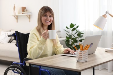 Woman in wheelchair with cup of drink using laptop at table in home office