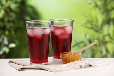 Photo of Refreshing hibiscus tea with ice cubes in glasses and honey on white wooden table against blurred green background