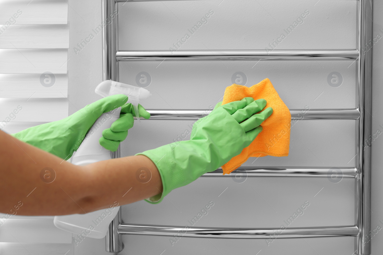 Photo of Woman cleaning heated towel rail with sprayer and rag, closeup