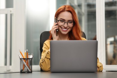Photo of Happy woman talking on smartphone while working with laptop at desk in office