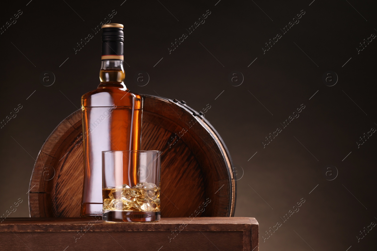 Photo of Whiskey with ice cubes in glass and bottle on wooden table near barrel against dark background, space for text