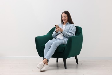 Photo of Beautiful woman using smartphone while sitting in armchair near white wall indoors