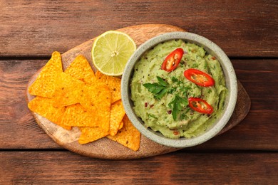 Photo of Bowl of delicious guacamole, lime and nachos chips on wooden table, top view