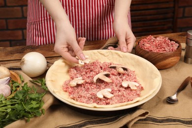 Photo of Woman putting mushrooms into baking dish with dough to make meat pie at wooden table, closeup
