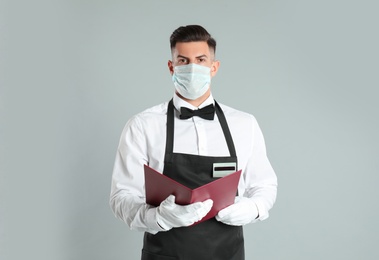 Photo of Waiter in medical face mask with menu on light grey background