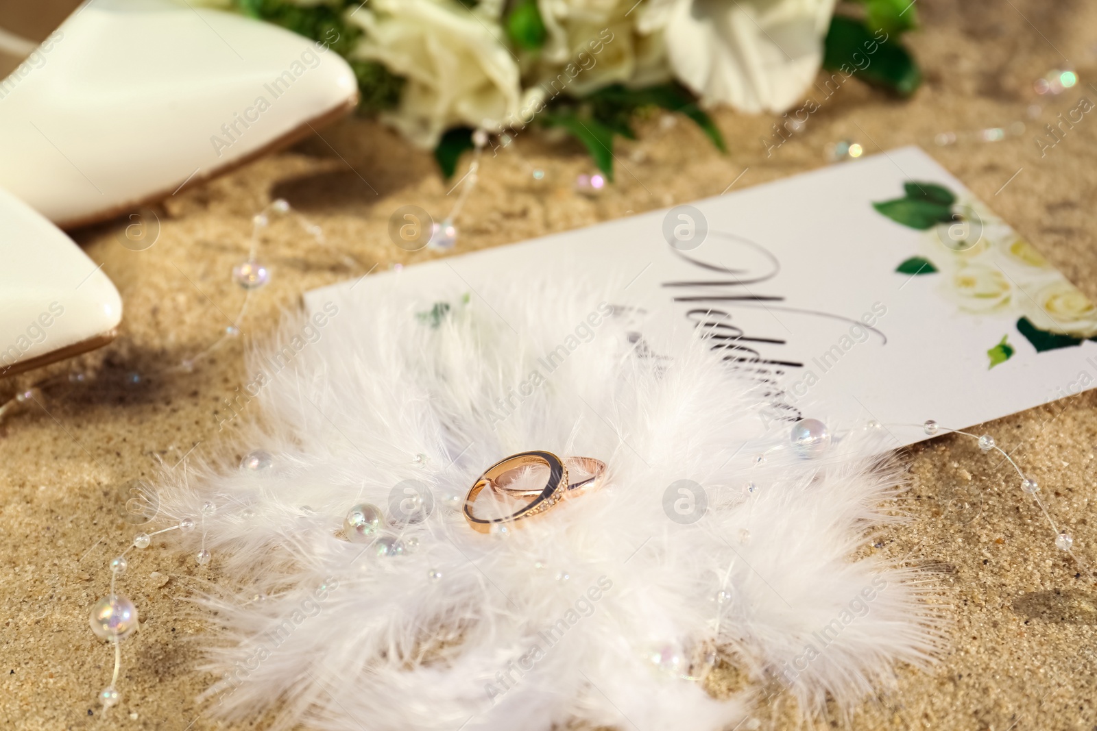 Photo of Gold wedding rings, invitation and decor on sandy beach, closeup