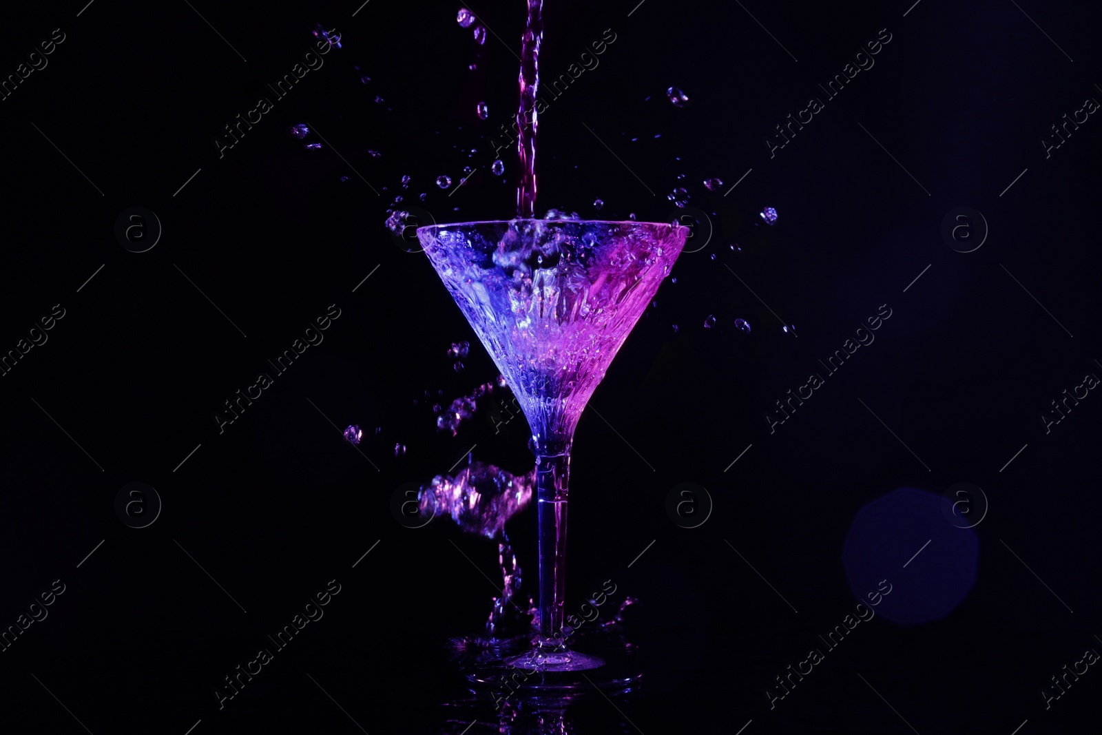 Photo of Pouring martini. Glass and splashes in neon lights on dark background, bokeh effect