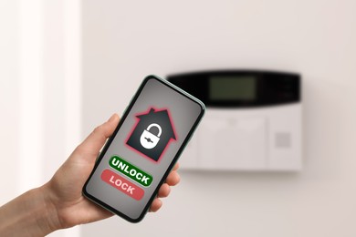 Woman operating home alarm system via mobile phone against white wall with security control panel, closeup. Application interface on device screen