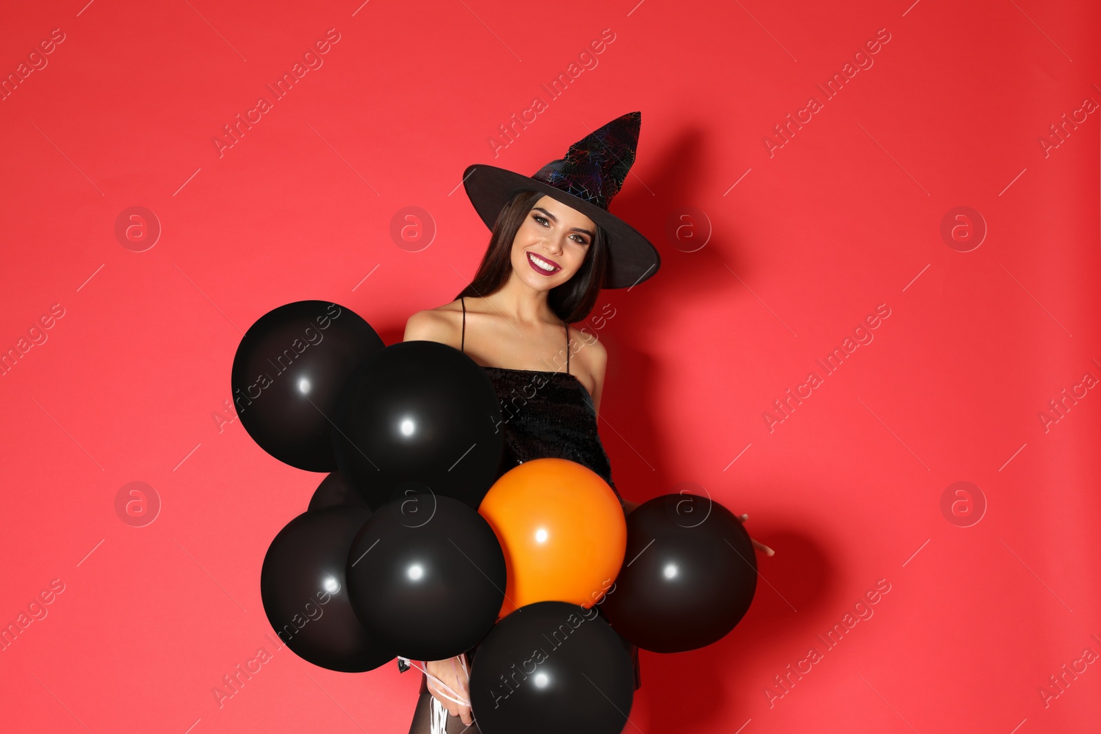 Photo of Beautiful young woman wearing witch costume with balloons for Halloween party on red background