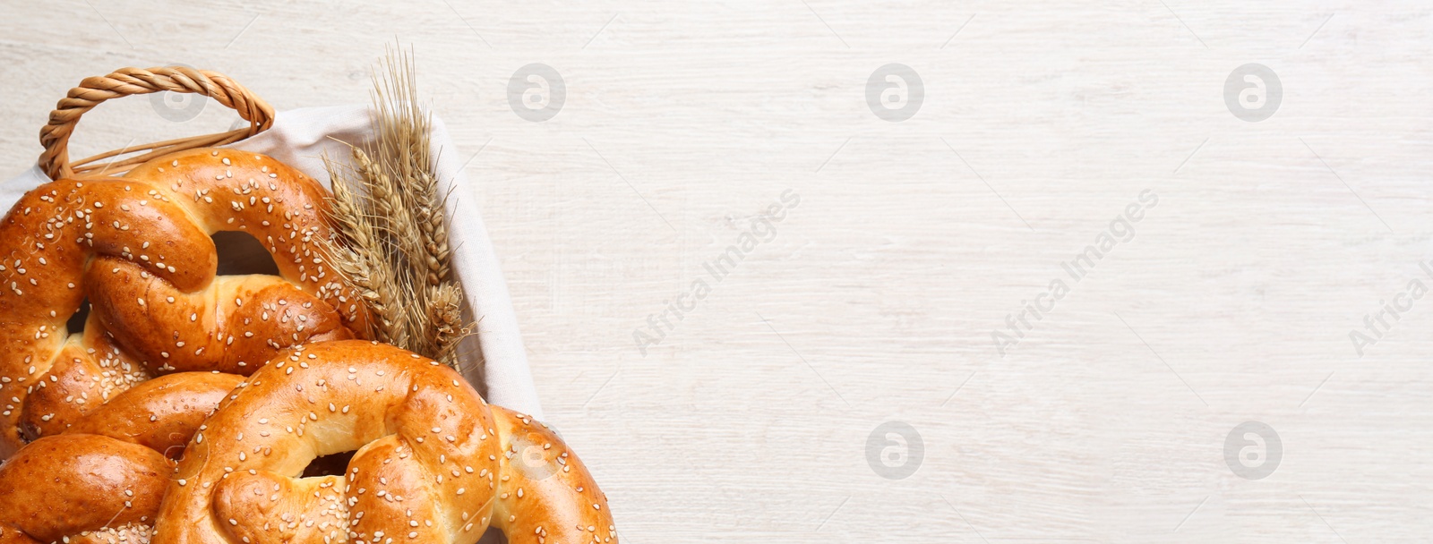 Image of Basket with delicious pretzels and wheat spikes on white wooden table, top view with space for text. Banner design