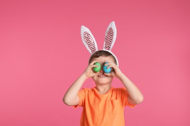 Photo of Little boy in bunny ears headband holding Easter eggs near eyes on color background