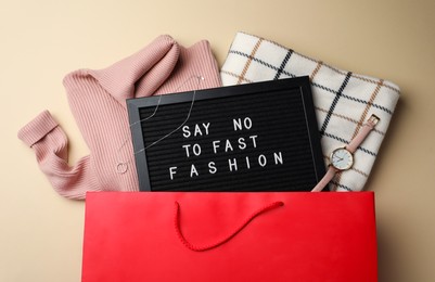 Photo of Black letter board with phrase SAY NO TO FAST FASHION, clothes and wristwatch in red shopping bag on beige background, flat lay