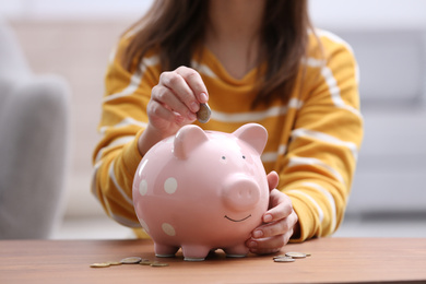 Woman putting coin into piggy bank at wooden table, closeup