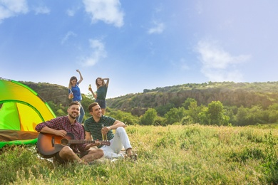 Photo of Group of young people resting with beer and guitar near camping tent in wilderness