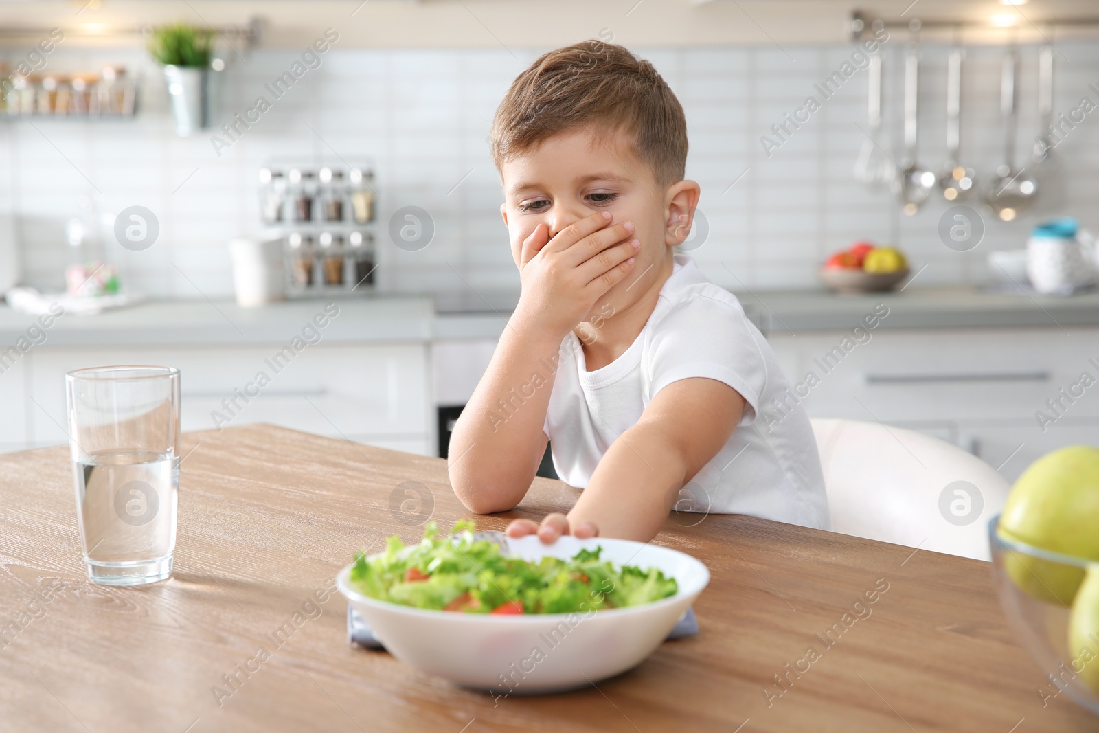 Photo of Little boy covering his mouth and refusing to eat vegetable salad at table in kitchen
