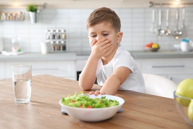 Photo of Little boy covering his mouth and refusing to eat vegetable salad at table in kitchen