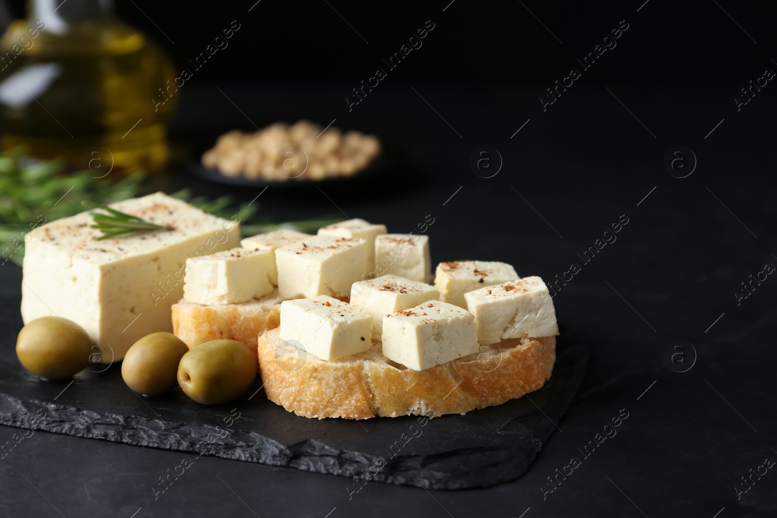 Photo of Pieces of delicious tofu with bread, rosemary and olives on black table. Soybean curd