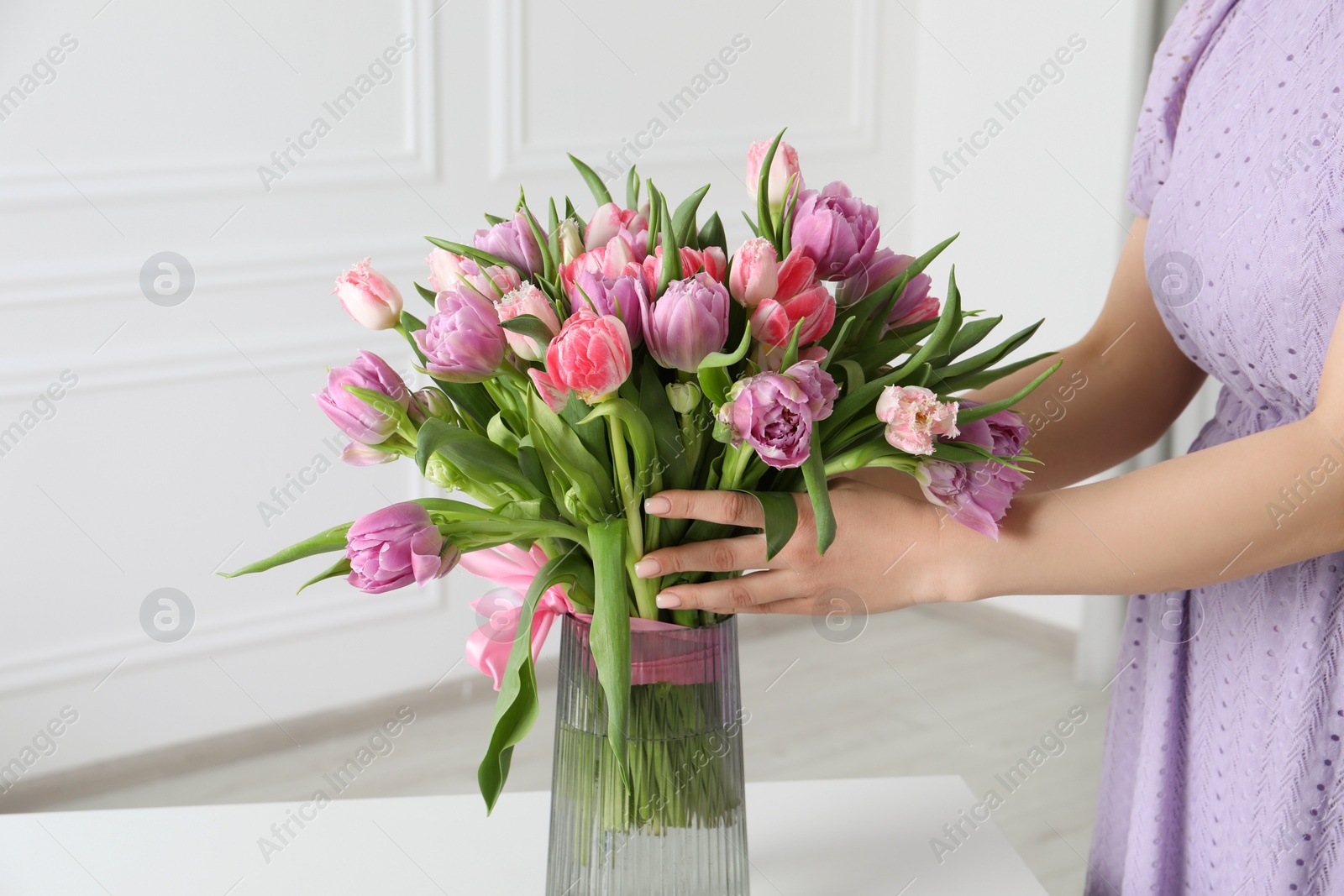 Photo of Woman putting bouquet of beautiful tulips in vase on white table indoors, closeup