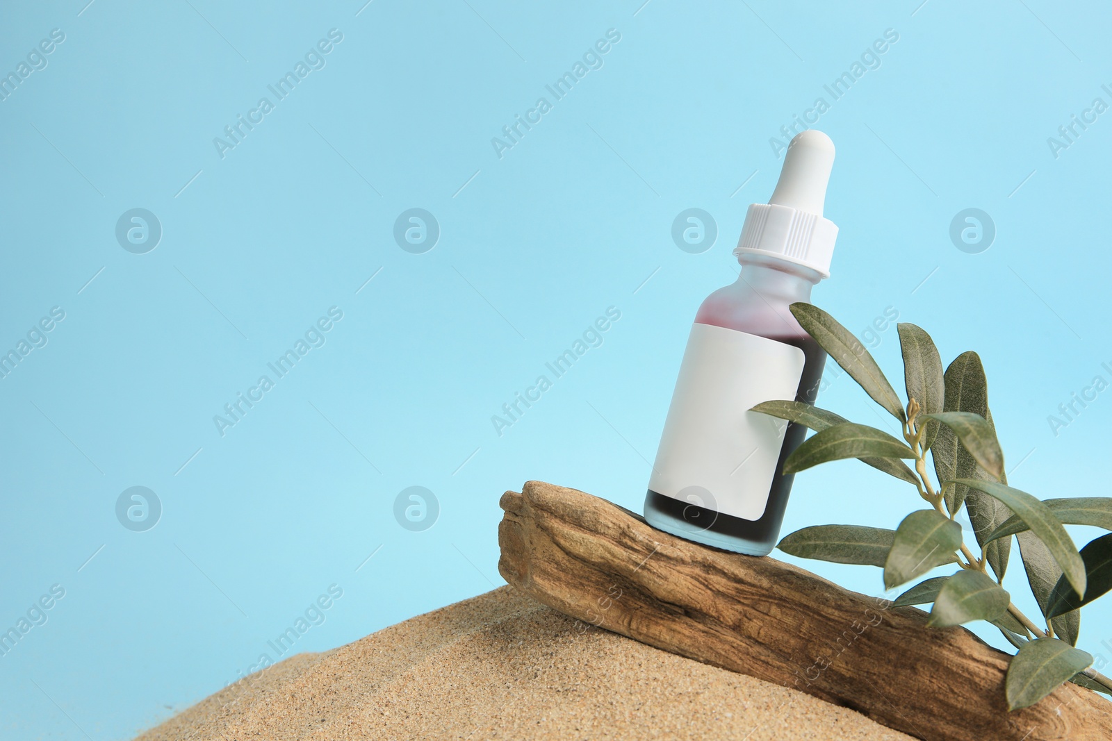 Photo of Bottle with serum, olive twig and bark on sand against light blue background, space for text. Cosmetic product