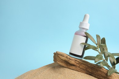 Bottle with serum, olive twig and bark on sand against light blue background, space for text. Cosmetic product